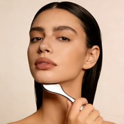 A woman with long, dark hair gently holds a silver facial tool against her jawline, showcasing her natural beauty.