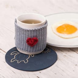 A round soft diatomaceous coaster in dark blue, supporting a cup with a knitted grey sleeve and a heart embellishment.
