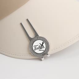 A silver golf divot tool with a ball marker attached, resting on a beige cap. It features a logo design.
