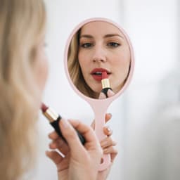 A pink handheld makeup mirror reflects a person applying lipstick, with a soft, neutral background.