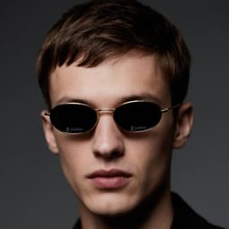 Oval retro sunglasses with a gold frame and dark lenses, worn by a model with short hair against a dark background.