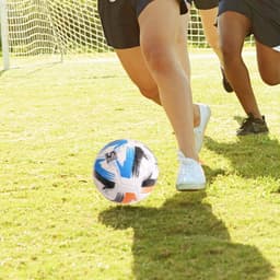 A promotional soccer ball with blue, black, and white patterns on a grassy field, alongside people playing soccer.