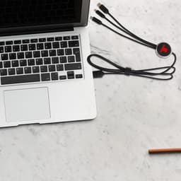 A black 3-in-1 charging cable with multiple connectors rests beside a laptop on a marble surface.