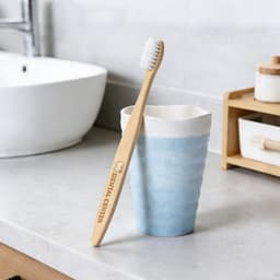 A large bamboo toothbrush with a light wooden handle and white bristles rests against a blue ceramic cup on a countertop.
