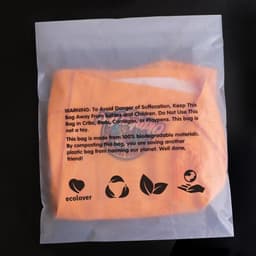 An eco-friendly biodegradable self-adhesive bag in a transparent plastic packaging, featuring an orange bag inside.