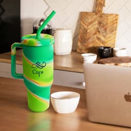 A drink bottle featuring a custom green and yellow silicone design with a handle and straw, alongside a laptop and bowls.