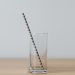 A drinking glass containing a stainless steel straw, placed on a wooden surface against a light background.
