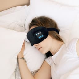 A pair of black Bluetooth sleep headphones worn by a person resting on a bed with white pillows.