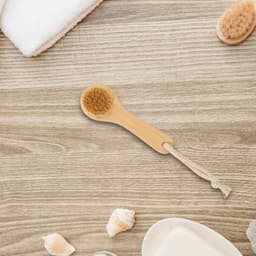 A natural wooden face brush with a beige bristle head, set against a wooden background with soft towels and seashells.