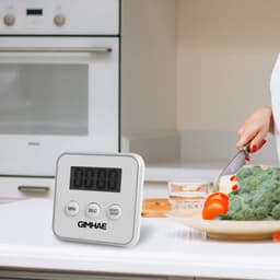A digital kitchen timer in white with a large display, placed on a kitchen countertop beside fresh vegetables.