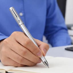 A silver 2 in 1 banner pen held in a hand, poised above a notebook on a desktop.