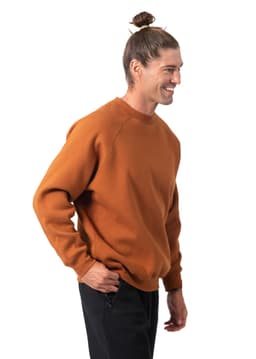 A man in a brown sweatshirt stands smiling, showcasing a casual style. The sweatshirt has a logo on it.