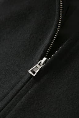 A close-up of a black wool blend varsity jacket featuring a silver zipper detail. The jacket has a logo.
