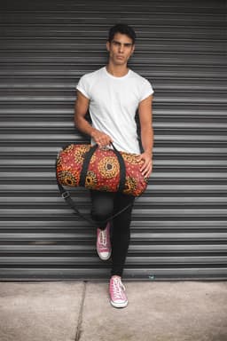 A duffle bag in red and black with a floral pattern is held by a person in a fitted white t-shirt and pink sneakers.
