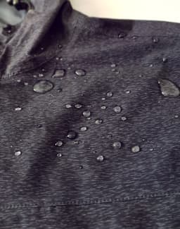 A close-up of a dark grey waterproof jacket with water droplets on the fabric and a hood. The jacket has a logo.