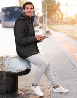 A man wearing a black unisex sustainable parka jacket, seated by a waterfront, holding a phone.