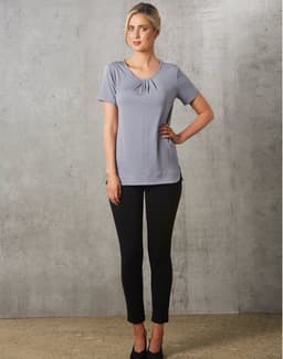 A grey short-sleeve knit top with pleats, paired with black fitted pants, worn by a model against a textured background.