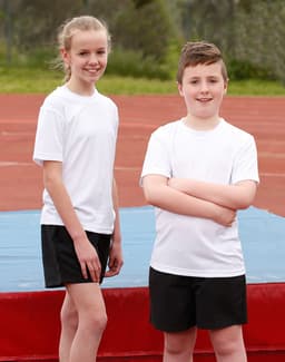 Two children wearing white short sleeve t-shirts and black shorts stand on a sports field. One child has their arms crossed.