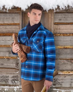 A man stands outdoors in a blue and black quilted flannel shirt, holding firewood near a wooden cabin.