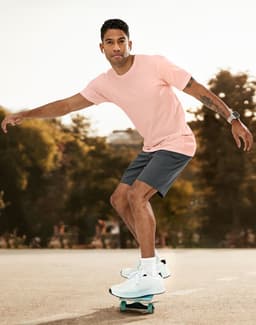 A man in a light pink premium cotton tee skates on a skateboard, wearing grey shorts and white sneakers.