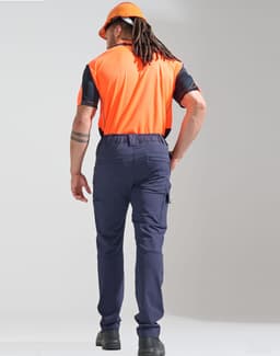 A man wearing orange and black work attire stands with his back to the camera, sporting navy blue flexi work pants and a helmet.