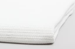 A white Cotton Cellular Blanket folded neatly, showcasing its texture and stitching. It features a branding logo.