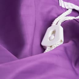 A purple laundry bag made of 100% polyester with a white drawstring and toggle closure. It features a textured surface.