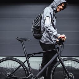A person in a grey rain jacket stands beside a black bicycle, wearing a backpack and smiling at the camera.