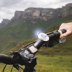 A bike handlebar with a clear light attachment and a digital display, set against a mountainous landscape.