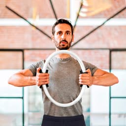 A man in a grey shirt holds a circular exercise ring in front of him, with a modern indoor backdrop.