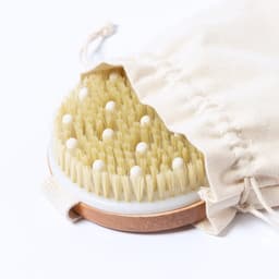 A round exfoliating massage brush with natural bristles and white nodules, resting in a cream-coloured pouch.