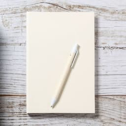 A cream notebook with a white Roliok pen resting atop it on a rustic wooden surface.