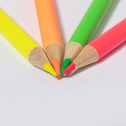 Four brightly coloured wooden pencils in yellow, orange, pink, and green, with pointed tips, arranged in a circle.
