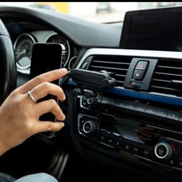 A car phone holder in black attached to a vehicle’s dashboard, with a hand placing a smartphone into it.
