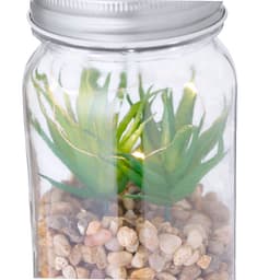 A glass jar containing faux green plants atop a bed of small decorative pebbles, with a metallic lid.