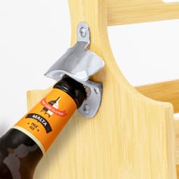 A drink bottle being opened with a metal bottle opener attached to a wooden surface. The bottle has an orange label.