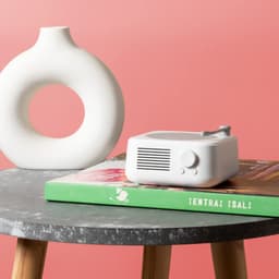 A Bluetooth speaker in a minimalist design, coloured white, sits on a stack of books next to a decorative vase on a table.