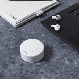 Earbuds in white beside a round charging case on a grey surface, with a notepad and laptop in the background.
