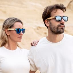 Sunglasses with blue lenses and wooden accents, worn by a man and woman against a sandy backdrop.