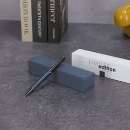 A metal pen in navy blue resting on a grey box, with a white box beside it featuring a logo.