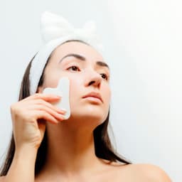 A woman using a white jade facial tool on her cheek, wearing a white hairband.
