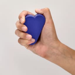 A purple heart-shaped stress ball being squeezed in a hand, featuring a textured surface.