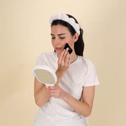 A woman wearing a white headband holds a mirror while applying makeup with a sponge. The background is a light colour.