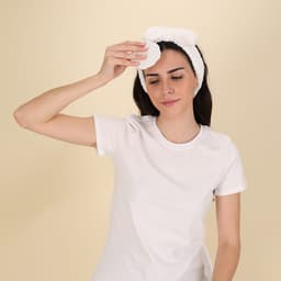 A white headband with a bow detail worn by a woman in a plain white t-shirt, applying a product to her face.
