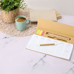 A gold planner with sticky notes, a ruler, a pen, a green coffee mug, and a potted plant on a marble surface.