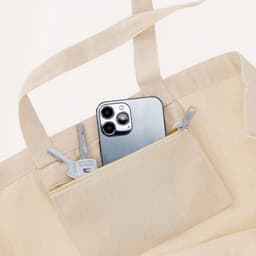 A beige tote bag with a smartphone and keys visible in the zipped pocket. The bag has sturdy handles and a logo.