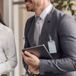 A man in a grey suit holds a tablet, standing next to a woman. Both are in a professional setting.