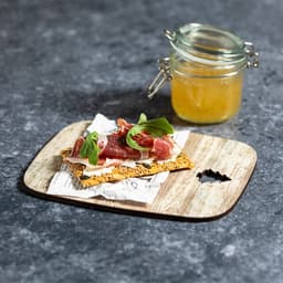 A wooden cutting board featuring a slice of crispbread topped with meat, herbs, and a small jar of preserves.