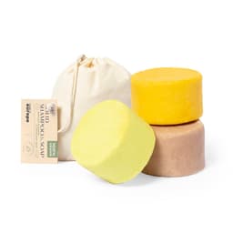 Three solid shampoo bars in yellow, brown, and cream, accompanied by a beige cloth pouch, with a product label.