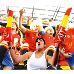 A woman cheerfully holds two red and yellow inflatable sticks in a crowd of supporters wearing red jerseys.